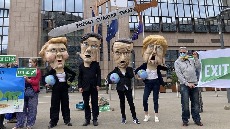 a mock sword with "Energy Charter Treaty" written on it hangs over the heads of protestors wearing masks of European leaders